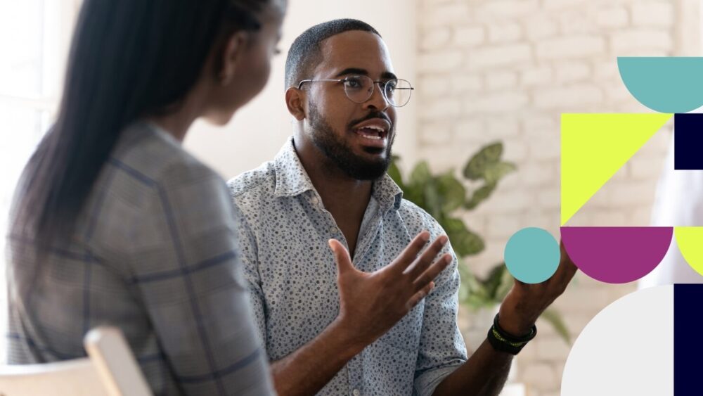 Two people discussing something, with a white wall and a plant in the background.